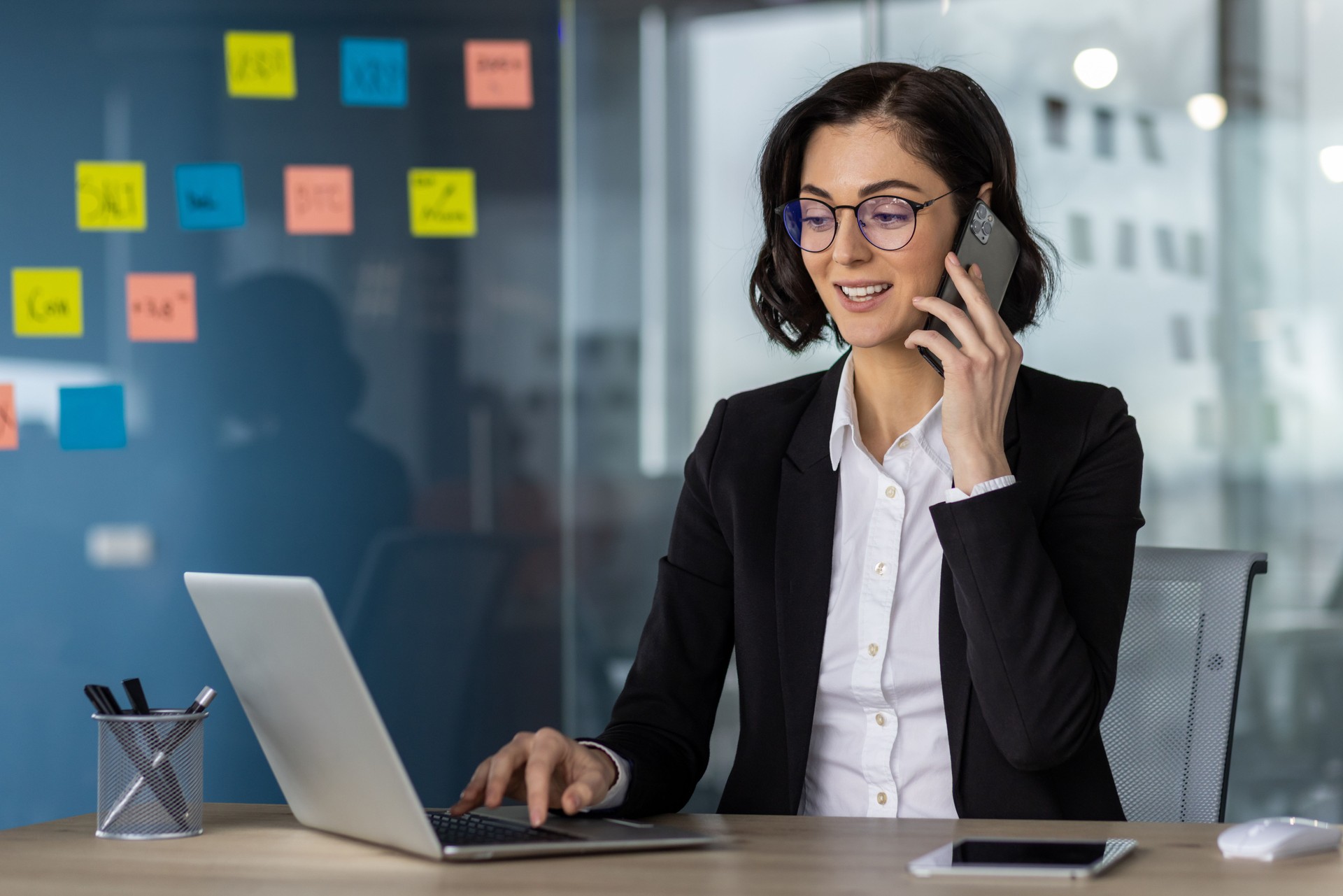 Businesswoman working in modern office on laptop and phone call, professional environment with sticky notes